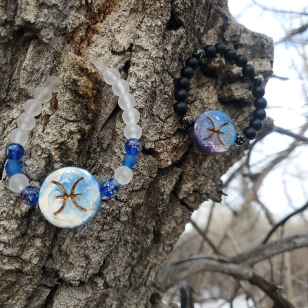 Two bracelets with Pisces Zodiac sign charms, hanging from a tree branch.